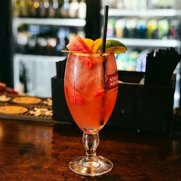 A refreshing red cocktail with watermelon and lime garnish sits on a wooden bar top.