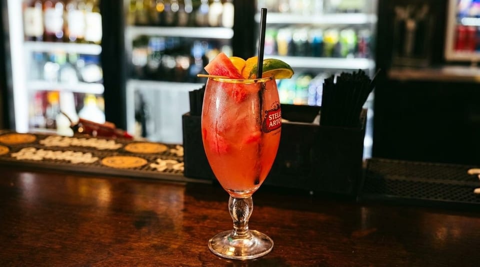 A refreshing red cocktail with watermelon and lime garnish sits on a wooden bar top.