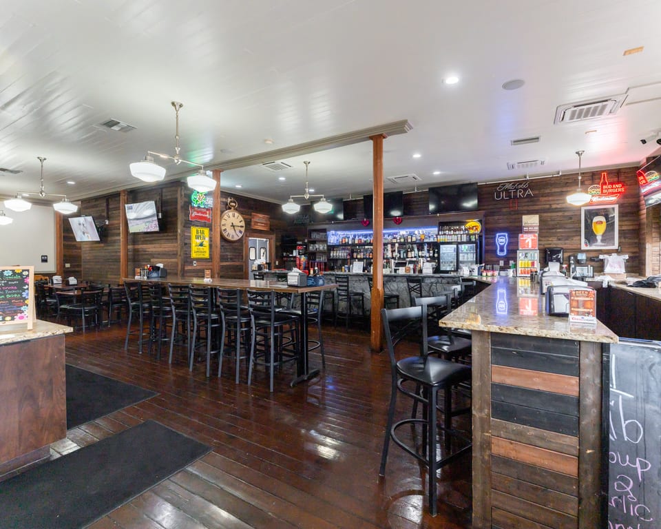 Interior of a bar with wooden walls and floors, featuring a long bar, high-top tables, and stools.