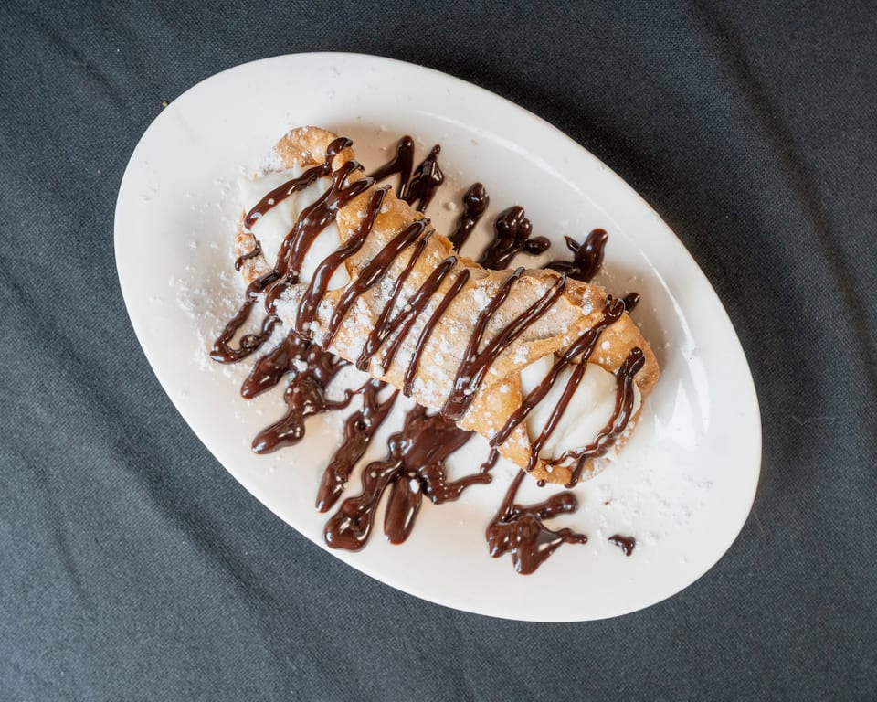 A cannoli drizzled with chocolate sauce and powdered sugar on a white oval plate.