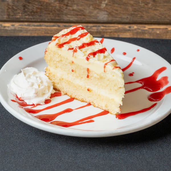 Slice of white cake with cream cheese frosting, strawberry drizzle, and whipped cream on a white plate.
