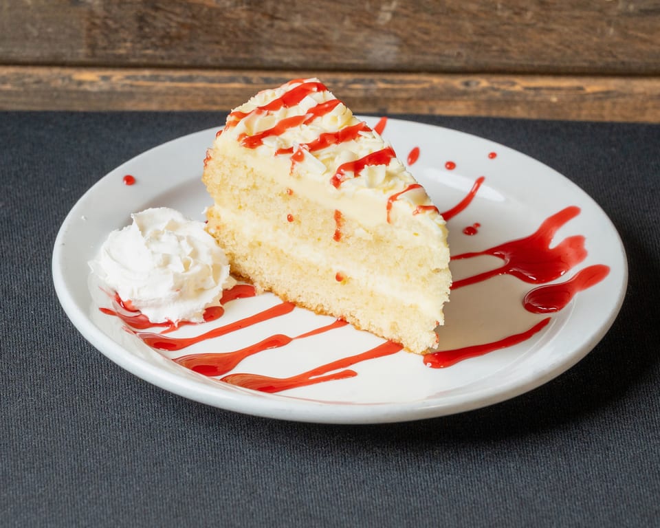 Slice of white cake with cream cheese frosting, strawberry drizzle, and whipped cream on a white plate.