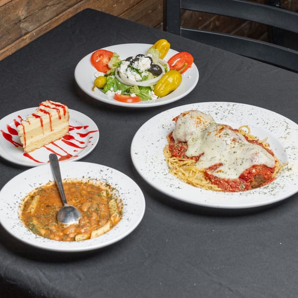 A table set with a Greek salad, spaghetti and meatballs, pasta fagioli, and cheesecake.