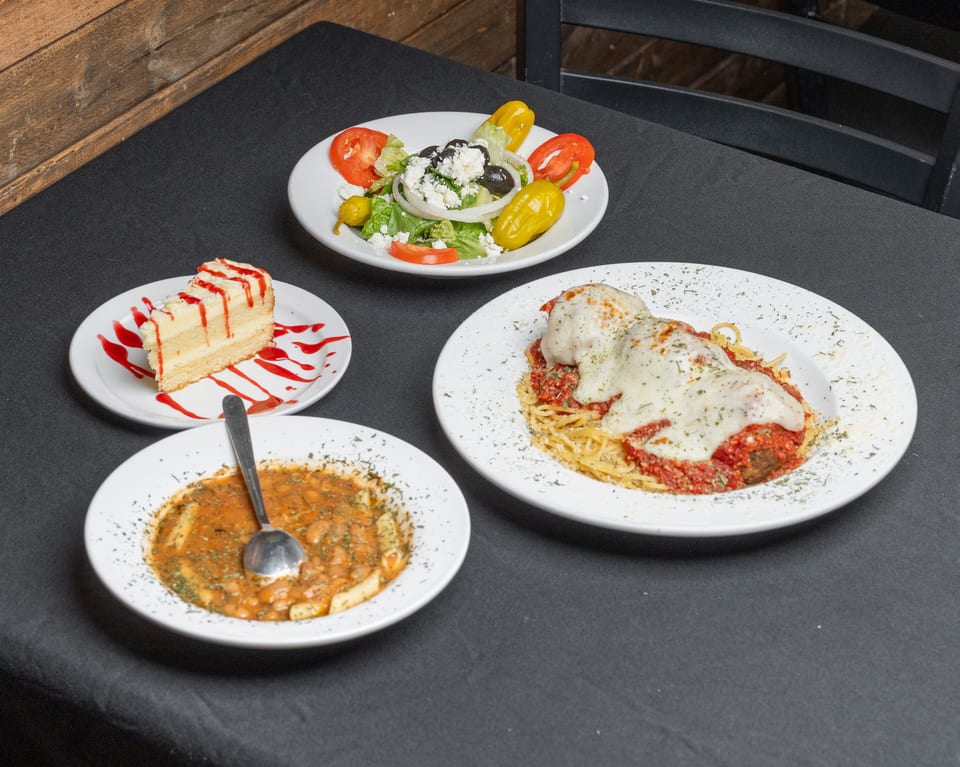 A table set with a Greek salad, spaghetti and meatballs, pasta fagioli, and cheesecake.