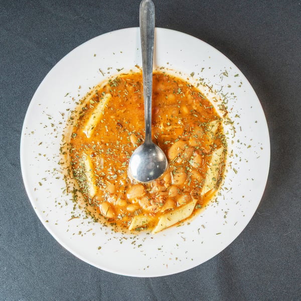 A white bowl of bean and pasta soup with a spoon, garnished with herbs.