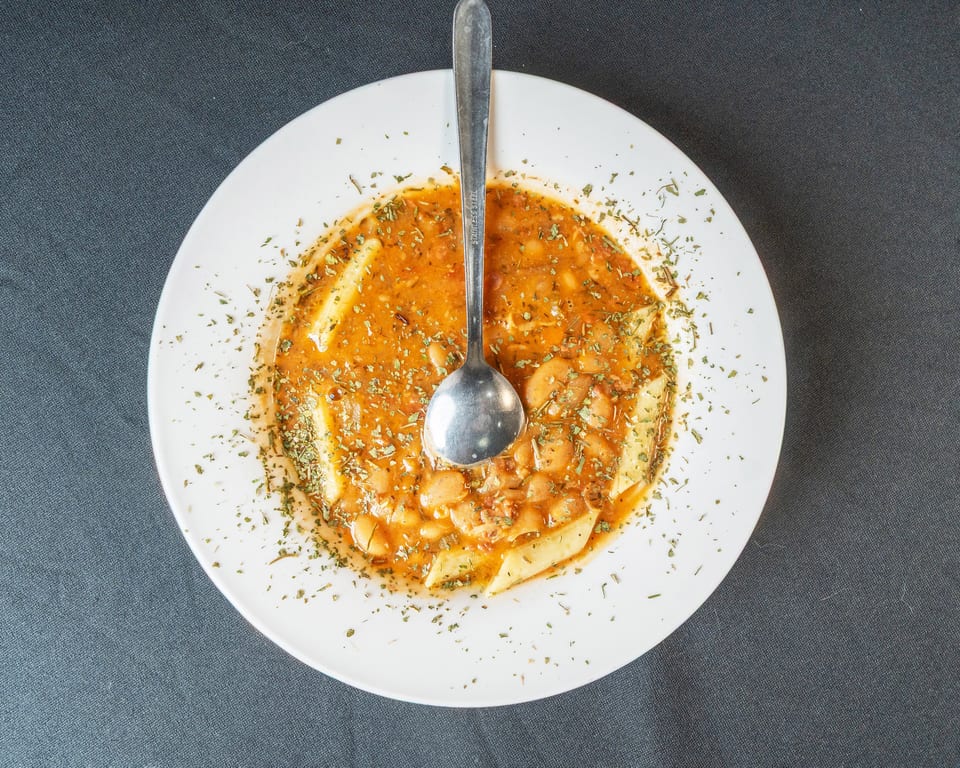 A white bowl of bean and pasta soup with a spoon, garnished with herbs.